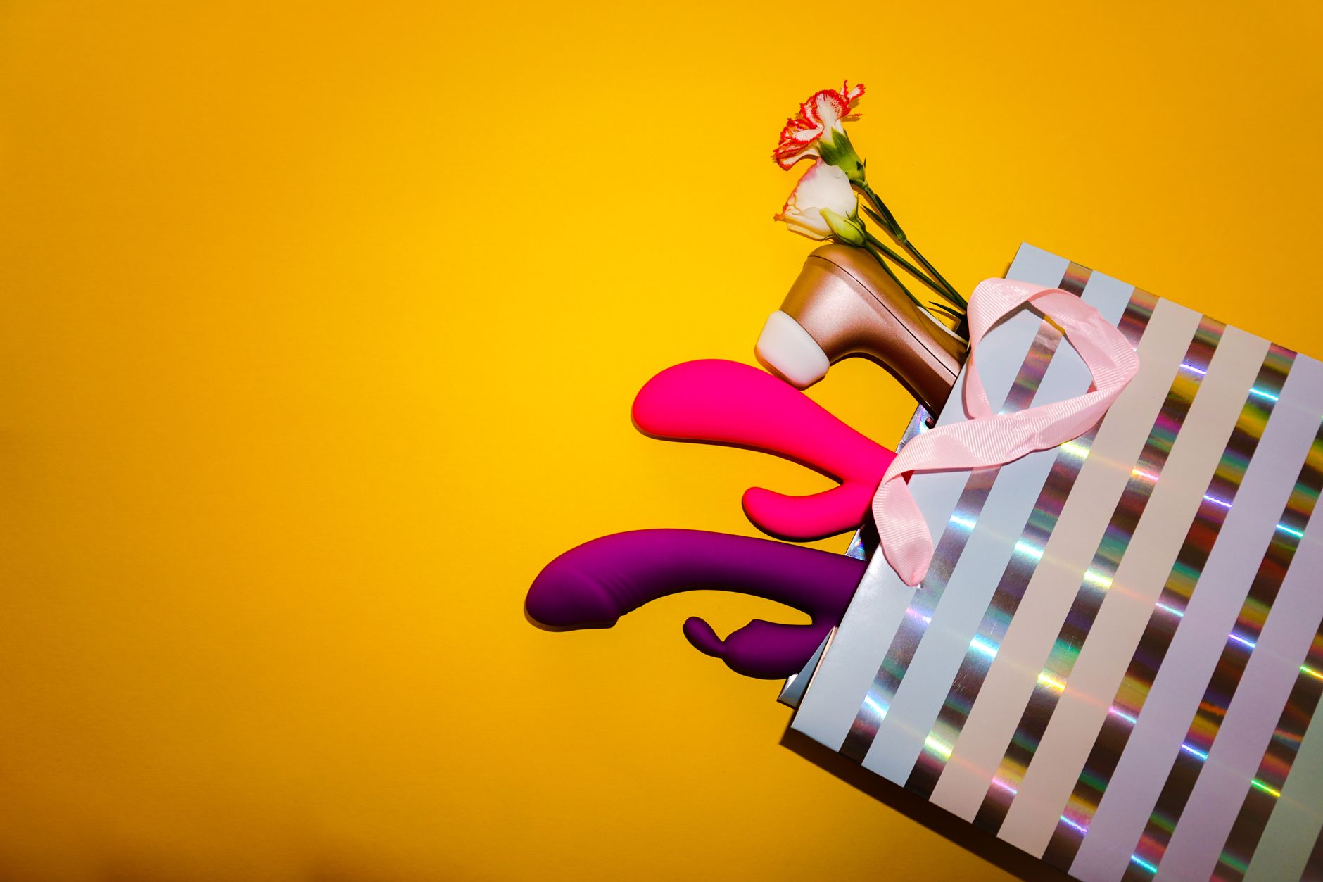Vibradores coloridos con flores en una bolsa de regalo holográfica sobre fondo amarillo vibrante