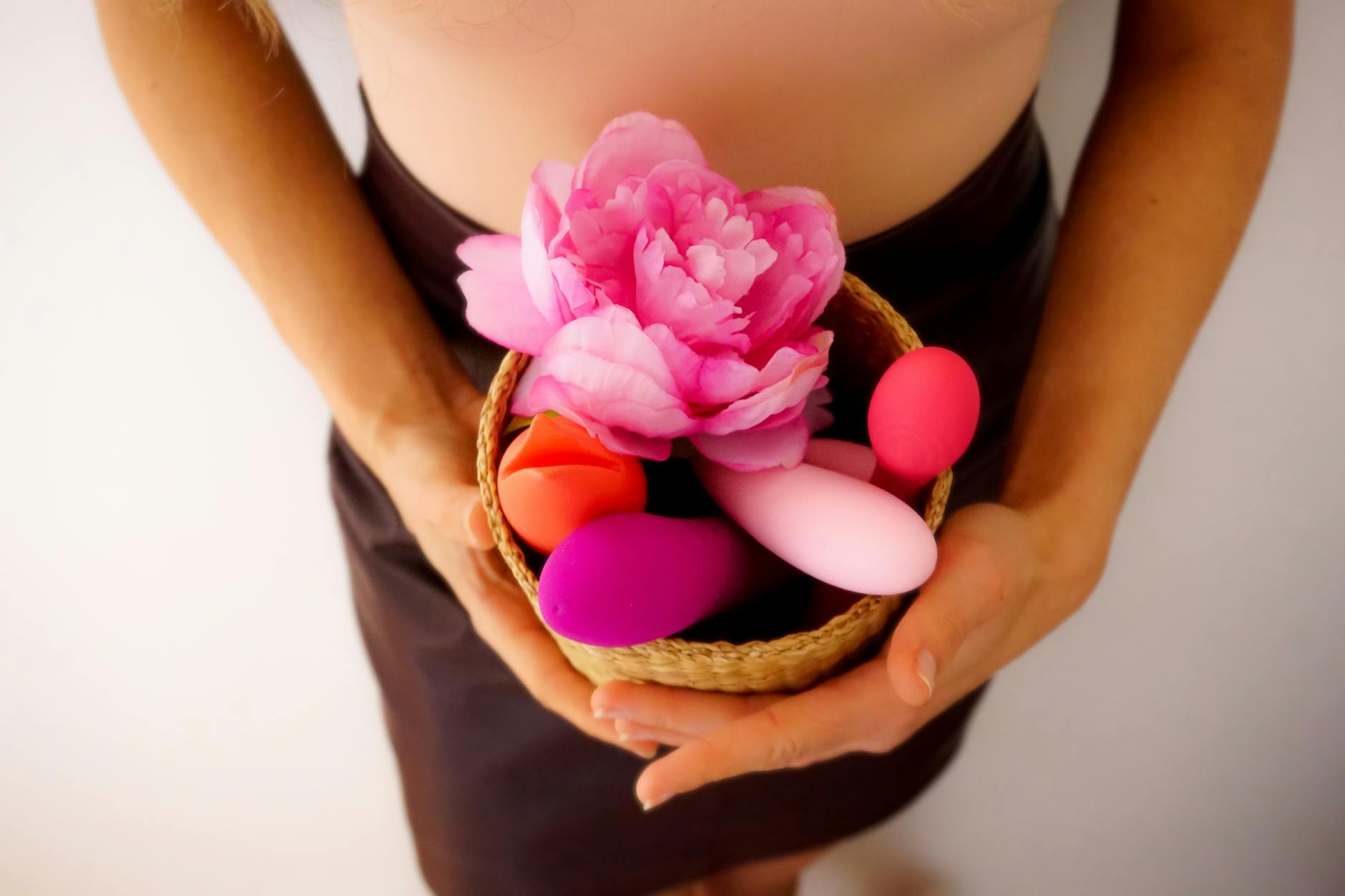 Una persona sostiene una canasta con vibradores coloridos y una flor rosa, representando el placer y la intimidad postparto.